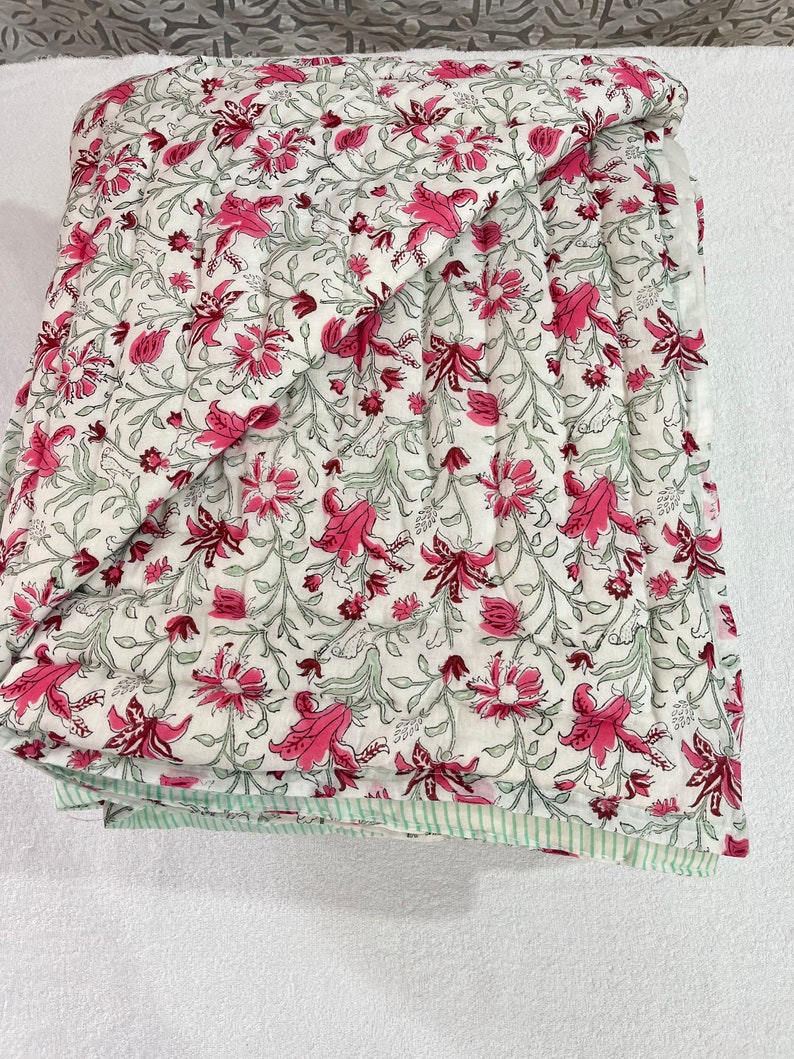 May include: A white cotton quilt with a pink floral pattern. The quilt is folded in half and has a green and white striped border.