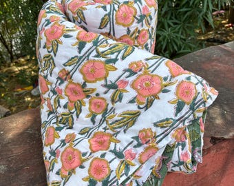 May include: Rolled-up floral quilt featuring a white background with a repeating pattern of pink flowers, yellow accents, and green leaves. The quilt's edges are a solid green color. The quilt is made of a soft, lightweight material.