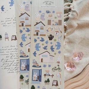 May include: A sheet of colorful journal stickers featuring a light blue, white, and brown color scheme. The stickers include illustrations of houses, birds, rabbits, stars, plants, a bicycle, a teapot, a cup of coffee, a slice of bread, and a sign that says "Rise and Shine".