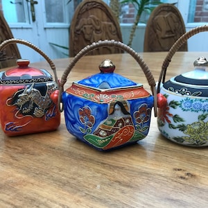 May include: Three small ceramic teapots with lids and bamboo handles. The teapots are decorated with colourful floral and dragon designs. The teapots are sitting on a wooden table.