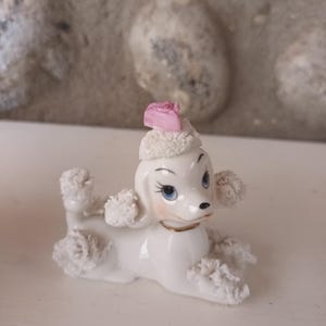 May include: A white ceramic poodle ornament with blue eyes, pink details, and a pink hat. The poodle has fluffy, textured details on its ears, paws, and tail. The figurine is lying down, with its legs extended.
