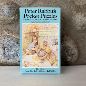 May include: A light blue box of "Peter Rabbit's Pocket Puzzles". The box features an illustration of Peter Rabbit and other characters from the story. The text on the box reads "12 Piece Wooden Jigsaw Puzzle, Hand cut by craftsmen".