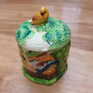 Może przedstawiać: Ceramiczny słoik z zielono-białą pokrywką, na której znajduje się żółty ptak. Słoik ma zielono-biało-brązowy wzór z małym domkiem z przodu.