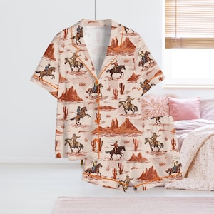 May include: A two-piece pajama set featuring a cowboy-themed print. The set includes a short-sleeved button-up shirt and matching shorts. The beige fabric is patterned with cowboys on horseback, cacti, and desert landscapes, outlined in orange.