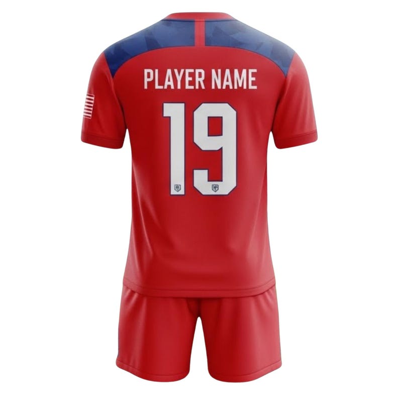 May include: Red soccer jersey and shorts set. The jersey has a blue and red design on the shoulders and the text "PLAYER NAME" above the number "19". The American flag is on the left sleeve.