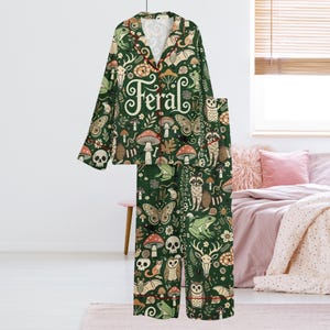 May include: Green pajama set with a whimsical forest pattern featuring mushrooms, butterflies, raccoons, and the word "Feral". The long-sleeved top has a notched collar and red trim. The matching pants complete the set.