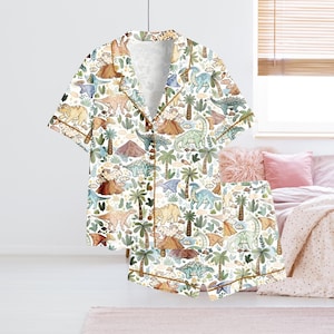 May include: White pajama set with a dinosaur pattern. The short-sleeved top and shorts feature a repeating design of various dinosaurs, palm trees, and volcanoes in shades of green, brown, and blue. The set has brown trim and buttons.