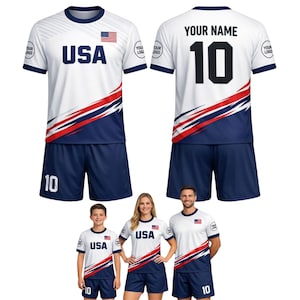 May include: White and navy blue USA soccer uniform set. The jersey has the word "USA" in large blue letters, a red and blue diagonal stripe design, and the number 10. The shorts are navy blue. The back of the jersey has the number 10 and the text "YOUR NAME".