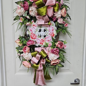 May include: A floral wreath with pink and red flowers, green leaves, and pink and green bows. A sign with the word "Hello" is in the center. The wreath is hanging on a white door.