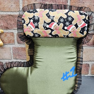 May include: A Christmas stocking-shaped pillow with a green body and a top section featuring gingerbread men wearing Santa hats and scarves on a black background. The pillow is trimmed with black lace and has the number 1 in blue.