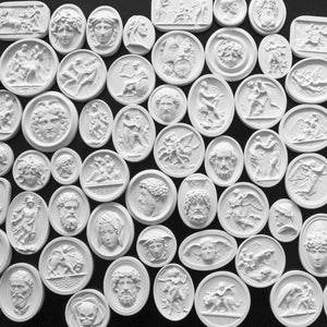 May include: A collection of white plaster relief sculptures, featuring classical figures and scenes. The oval and rectangular shapes showcase detailed depictions of faces, figures, and mythological scenes. The sculptures are arranged on a dark surface.