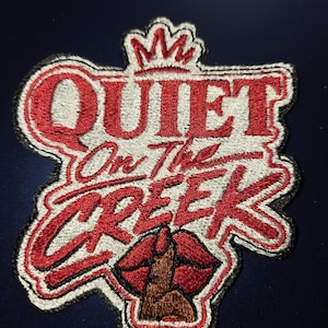 May include: A fabric patch featuring the red embroidered text "QUIET On The CREEK" with a crown icon above the word "QUIET". Below the text is an embroidered graphic of red lips with a brown finger pressed against them in a shushing gesture.