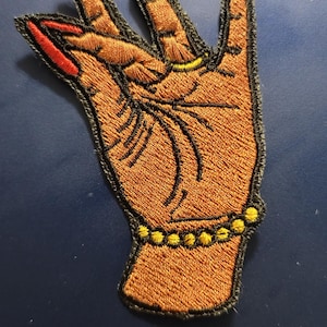 Może przedstawiać: Haftowana naszywka przedstawiająca dłoń w geście 'hang loose', z brązowym odcieniem skóry, czerwonymi paznokciami i żółtą bransoletką z koralików. Naszywka ma czarną obwódkę.