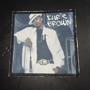May include: Square patch featuring a photo of Chris Brown in a white jacket and hat, holding a microphone. The text "CHRIS BROWN" is printed in white on the top right corner. The patch has a black border.