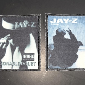 Könnte beinhalten: Zwei quadratische Aufnäher mit Albumcovern. Der linke Aufnäher zeigt eine Figur mit Hut, die eine Zigarre raucht, mit dem Text "JAY-Z" und "REASONABLE DOUBT". Der rechte Aufnäher zeigt eine rauchende Figur mit "JAY-Z" und "IN MY LIFETIME".