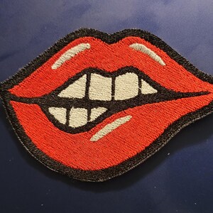 Può includere: Toppa ricamata raffigurante una bocca rossa stilizzata con denti bianchi e un contorno nero. La toppa ha un aspetto strutturato, con le labbra rosse e i denti bianchi che risaltano sul bordo nero. Adatta per abbigliamento o accessori.