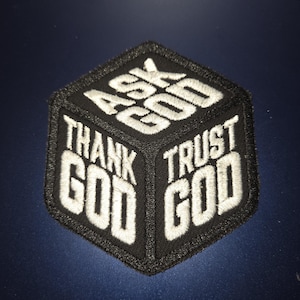 May include: A black hexagonal embroidered patch featuring white stitched text arranged in a cube-like perspective. The top section reads ASK GOD, the left side reads THANK GOD, and the right side reads TRUST GOD, all in a bold, sans-serif font.