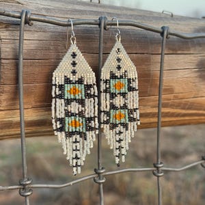 May include: Beaded earrings with a geometric pattern. The earrings have a cream base with black, turquoise, and orange details. They hang from silver-coloured hooks. The earrings are shown against a wooden fence.