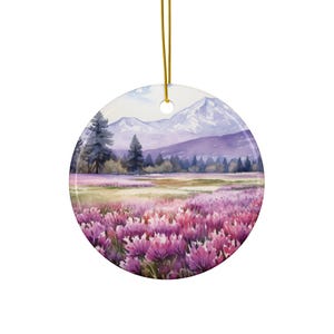 May include: A watercolor painting of a mountain range with a field of purple flowers in the foreground. The mountains are snow-capped and the sky is a light blue. The painting is on a round ceramic ornament.