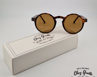 May include: Round tortoiseshell sunglasses with brown lenses. The sunglasses are sitting on a white box with the text "Chez Yvette" printed on it. The text says "Boot & Shoe Maker" and "For Gents & For Ladies" and "Accessories & Jewelry".