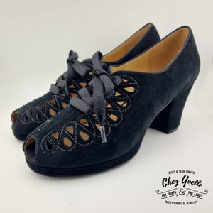 Può includere: Tacchi neri in pelle scamosciata con punta aperta, design a taglio e lacci in nastro nero. I tacchi sono alti circa 7,5 cm. Le scarpe sono realizzate da Chez Yvette, un produttore di stivali e scarpe per uomini e donne.