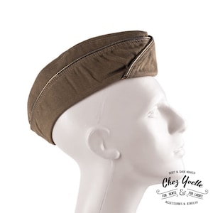 Op de afbeelding: Olijfgroene militaire pet met een zilveren ritssluiting langs de rand. De pet wordt getoond op een witte mannequinhoofd. Een accessoire in vintage stijl.