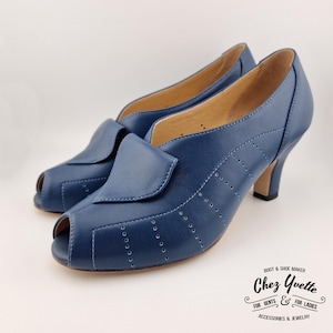 Può includere: Un paio di tacchi peep-toe in pelle blu. Le scarpe hanno un tacco piccolo e una patta sulla parte superiore del piede. Sono visibili cuciture decorative e perforazioni. Il marchio "Chez Yvette" è stampato in basso a destra.