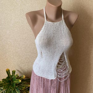 May include: Cream-coloured halter top with a knitted texture. The top features a unique design with a side slit and fringed edges. The garment is displayed on a mannequin, showcasing its shape and style. The top is paired with a pink skirt.