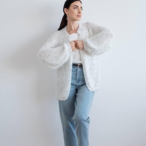 May include: A white open-front knit cardigan featuring long, voluminous sleeves and scattered silver sequins throughout the fabric. The garment is paired with a white top, light blue denim jeans with a brown belt, and light-colored woven flats.