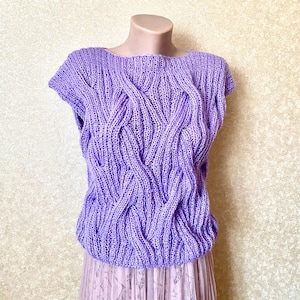Op de afbeelding: Een lavendelkleurige, mouwloze gebreide top met een textuur kabelbreisel ontwerp. De top wordt op een mannequin getoond en heeft een ronde halslijn. De onderkant van de top is licht gerimpeld. De top wordt gecombineerd met een lichtroze rok.
