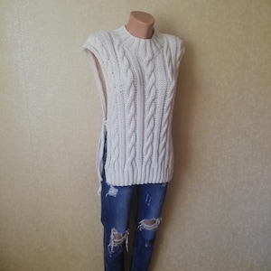 Könnte beinhalten: Cremefarbene Strickweste mit Rundhalsausschnitt und Seitenschlitzen. Das ärmellose Oberteil hat ein strukturiertes Zopfmuster und wird mit blauen Jeans mit Distressed-Details kombiniert.