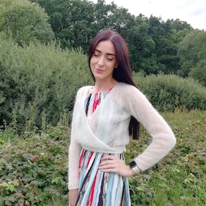 Könnte beinhalten: Ein hellbeige, langärmliger Wickelpullover. Der Pullover ist aus einem weichen, flauschigen Material gefertigt. Die Person trägt darunter ein farbenfrohes, gestreiftes Kleid. Der Hintergrund ist ein üppiges Grün mit Bäumen und Blättern.