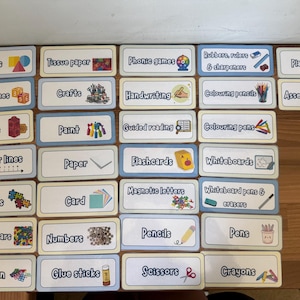 May include: A collection of rectangular classroom organisation labels with light blue borders. Each label features black text and a corresponding colourful icon representing school supplies like shapes, crafts, paint, paper, puzzles, cards, and various writing tools.