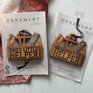 May include: Two wooden toolbox ornaments with tools inside. One says "DAD'S LITTLE HELPER" and the other says "GRANDPA'S LIL HELPER". Both are packaged in clear plastic with a black string for hanging. The brand name "Jellybeans Market" is printed on the packaging.