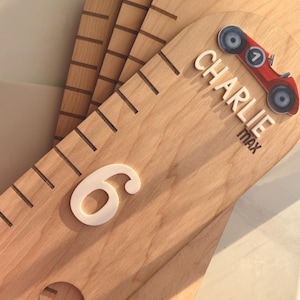 May include: A light-coloured wooden growth chart ruler featuring engraved measurement markings. The top section displays the name CHARLIE MAX in white lettering, accompanied by a small red toy car graphic and a large white number 6.