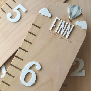 May include: A light-colored wooden growth chart featuring white raised numbers, including 5 and 6. The top is rounded and decorated with white clouds, a hot air balloon illustration, and the name FINN with the name beckett gerald below it.