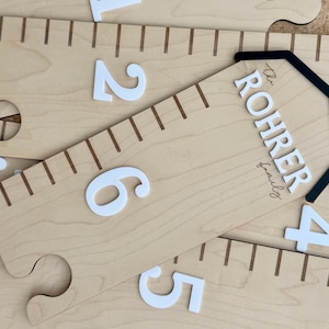 May include: A light-coloured wooden growth chart ruler featuring dark brown measurement markings and large white raised numbers. The top section displays the text "The ROHRER family" in white lettering beneath a black arched outline design.