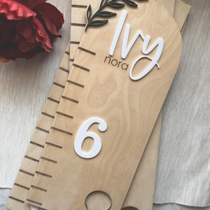 May include: Wooden growth chart with the name "Ivy" and the number "6" in white. The chart has a measuring scale and a decorative olive branch. The background is a light wood colour.