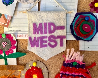 Könnte beinhalten: Ein Banner aus lila Filz mit den Worten "MID-WEST" auf weißem Hintergrund. Das Banner ist an einer Korktafel mit anderen Bastelartikeln befestigt, darunter eine rote Filzblume, eine gewebte Wandteppich und eine mehrfarbige Filzblume.