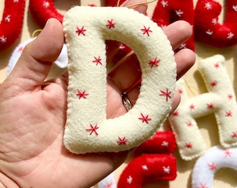 May include: A cream-colored felt letter D ornament featuring small red embroidered star-shaped accents. The letter has a thick, padded construction with visible stitching along the edges and a thin gold hanging loop attached to the top center.