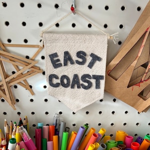 Könnte beinhalten: Ein weißes Stoffbanner mit den Worten "EAST COAST" in schwarzen Filzbuchstaben. Das Banner hängt an einer weißen Lochwand.