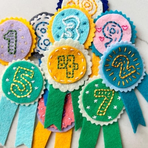 Peut inclure: Un ensemble de badges en feutre avec des numéros brodés de 1 à 9. Chaque badge a une couleur et un design différents. Les badges sont attachés à des rubans bleus, verts et jaunes.