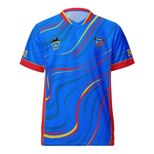 Könnte beinhalten: Blaues Trikot mit roten und gelben Akzenten. Es hat einen V-Ausschnitt und kurze Ärmel mit rotem Besatz. Das Trikot hat ein Wirbelmuster und enthält den Text "CONGO" und ein Schildemblem. Die Nummern 43 und 24 sind auf den Ärmeln sichtbar.