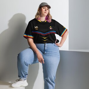 Puede incluir: Camiseta negra con dise&ntilde;o de cuello en V, con una corona y un emblema en el pecho. La camiseta tiene rayas rojas, amarillas y azules en las mangas y un dise&ntilde;o similar en el pecho. Se usa con vaqueros azul claro y una gorra gris.