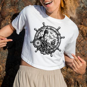 May include: White crop top featuring a black and white nautical design. The design includes a ship's wheel, a lighthouse, a compass, an anchor, and a sailboat. The shirt is made of a soft, comfortable material.