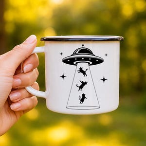 May include: A white enamel mug with a black rim featuring a black graphic of a UFO beaming up three small cats. The design includes four four-pointed stars surrounding the spacecraft. The mug has a curved handle on the left side of the body.