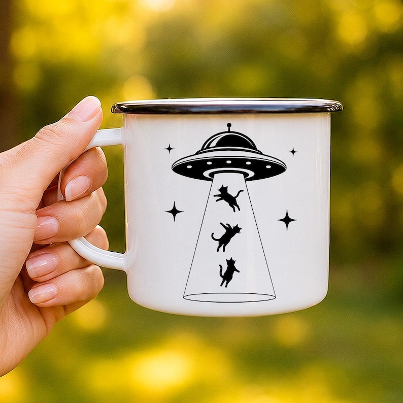 May include: A white enamel mug with a black rim featuring a black graphic of a UFO beaming up three small cats. The design includes four four-pointed stars surrounding the spacecraft. The mug has a curved handle on the left side of the body.