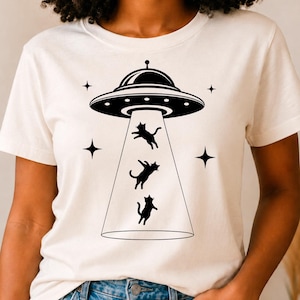 May include: A cream-colored short-sleeve t-shirt featuring a black graphic print of a flying saucer emitting a beam of light. Three black cat silhouettes are depicted floating within the beam, surrounded by four small black four-pointed stars.