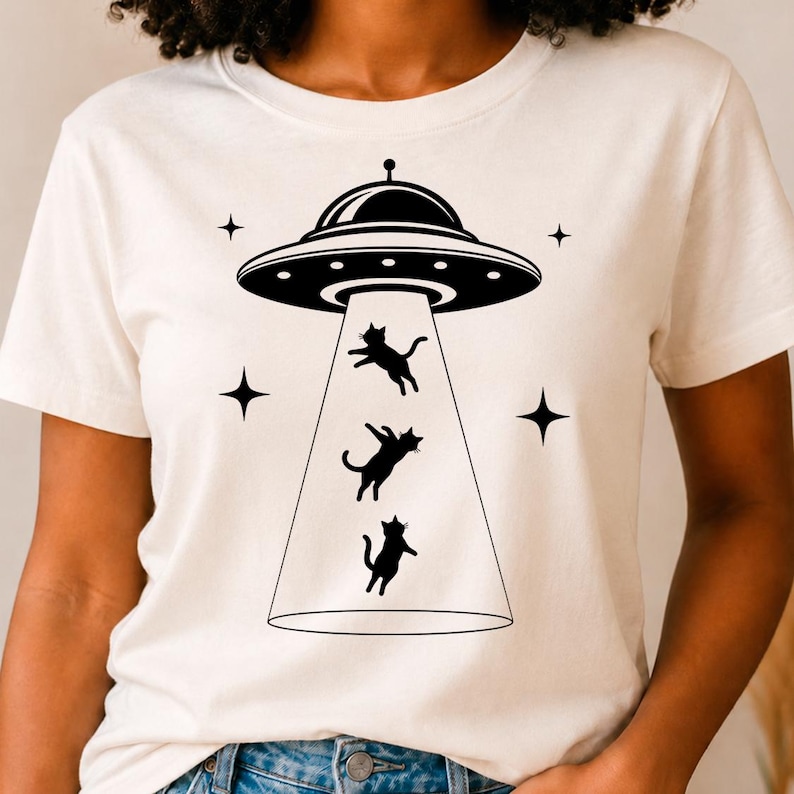 May include: A cream-colored short-sleeve t-shirt featuring a black graphic print of a flying saucer emitting a beam of light. Three black cat silhouettes are depicted floating within the beam, surrounded by four small black four-pointed stars.