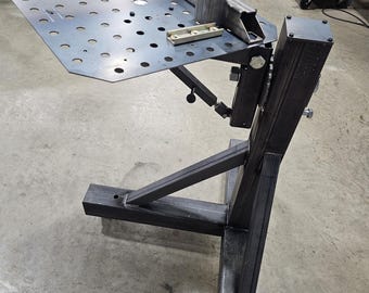 May include: A metal welding table with a perforated surface and adjustable components. The table features a dark gray finish and is designed for industrial use. It has a sturdy base and various clamps and supports.